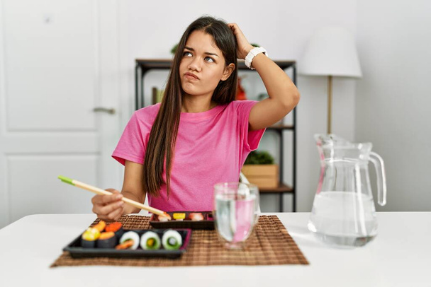 Mujer morena joven comiendo sushi usando palillos confundir y preguntarse acerca de la pregunta. incierto con la duda, pensando con la mano en la cabeza. concepto pensativo.  - Foto, Imagen