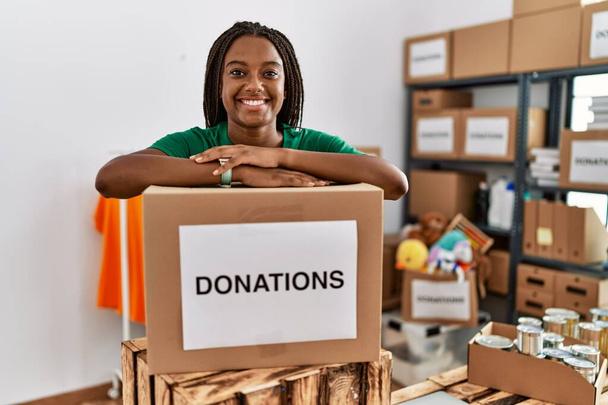Νεαρή Αφροαμερικανή που φοράει στολή εθελοντή και στηρίζεται σε πακέτο δωρεών στο φιλανθρωπικό κέντρο - Φωτογραφία, εικόνα