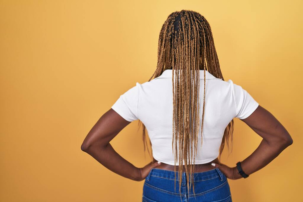 アフリカ系アメリカ人女性とともに編組髪立ち上の黄色の背景立って後ろに見ます離れて腕で体  - 写真・画像