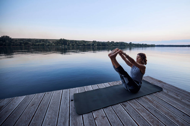 A fit yogi woman is practicing yoga on a dock at dusk. She is in the Boat yoga posture. - 写真・画像