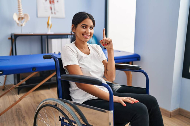 Fizyoterapi kliniğinde tekerlekli sandalyede oturan genç İspanyol kadın bir fikir veya soruyla gülümsüyor ve mutlu bir yüzle işaret ediyor.  - Fotoğraf, Görsel