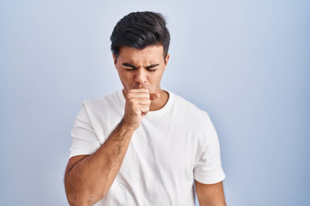 青い背景に立っているヒスパニック系の男性は具合が悪く、風邪や気管支炎の症状として咳をします。ヘルスケアの概念.  - 写真・画像