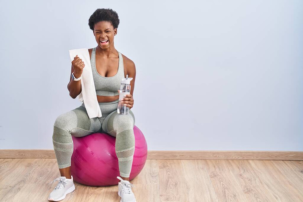 Femme afro-américaine portant des vêtements de sport assis sur pilates balle très heureux et excité de faire geste gagnant avec les bras levés, souriant et criant pour le succès. concept de célébration.  - Photo, image