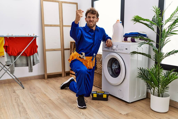 Young hispanic man working on washing machine annoyed and frustrated shouting with anger, yelling crazy with anger and hand raised  - Photo, image