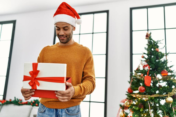 Hombre árabe joven sonriendo feliz unboxing regalo de pie junto al árbol de Navidad en casa. - Foto, Imagen