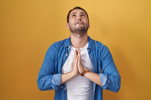 Latinomies seisoo keltaisen taustan päällä kerjäämässä ja rukoilemassa kädet yhdessä toivon ilme kasvoillaan hyvin emotionaalinen ja huolestunut. kerjääminen.  - Valokuva, kuva