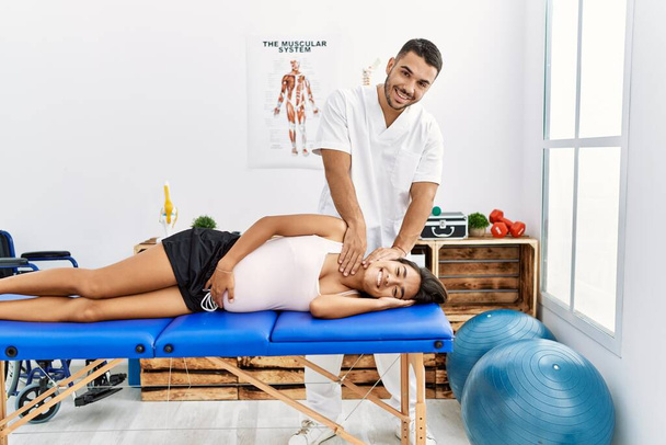 Latinský muž a žena v fyzioterapeutické uniformě s těhotenskou rehabilitační sezení natahování krku na fyzioterapeutické klinice - Fotografie, Obrázek