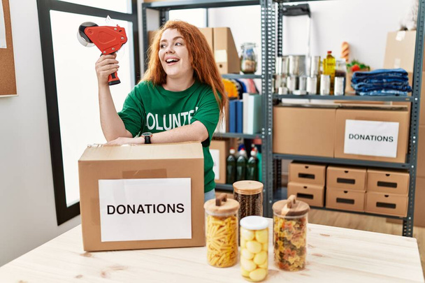 Νεαρή κοκκινομάλλα που φοράει στολή εθελοντή συσκευάζοντας κουτιά δωρεών στο φιλανθρωπικό κέντρο - Φωτογραφία, εικόνα