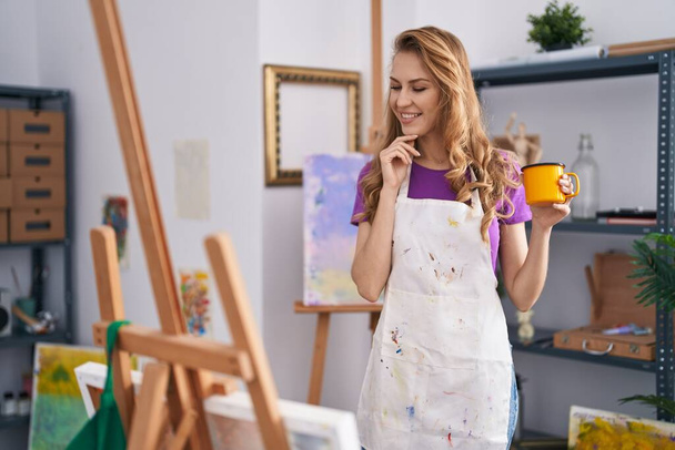 Молодая блондинка-художник, уверенно улыбающаяся, пьет кофе в художественной студии - Фото, изображение