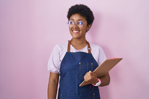 Młoda Afroamerykanka ubrana w profesjonalny fartuch kelnerski, trzymająca podkładkę, patrząca w bok z uśmiechem na twarzy, naturalnym wyrazem twarzy. śmiać się pewnie.  - Zdjęcie, obraz