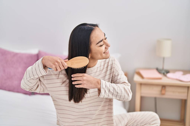 Giovane donna ispanica pettinando i capelli seduta sul letto in camera da letto - Foto, immagini