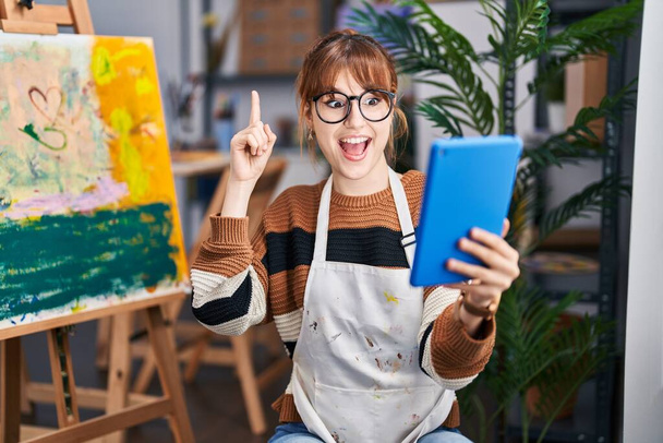 Νεαρή όμορφη καλλιτέχνης γυναίκα κάνει βιντεοκλήση με tablet χαμογελώντας με μια ιδέα ή ερώτηση δείχνοντας δάχτυλο με χαρούμενο πρόσωπο, νούμερο ένα  - Φωτογραφία, εικόνα
