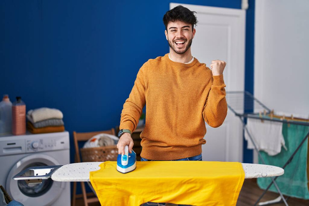 Hispanic man with beard ironing clothes at home screaming proud, celebrating victory and success very excited with raised arms  - Foto, Imagem