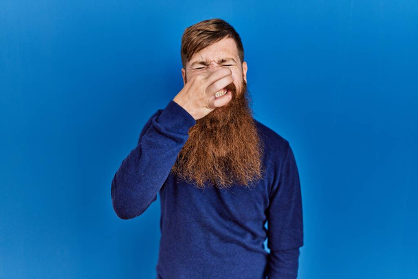 Homme rousse à longue barbe portant un pull bleu décontracté sur fond bleu sentant quelque chose de puant et dégoûtant, odeur intolérable, retenant la respiration avec les doigts sur le nez. mauvaise odeur  - Photo, image