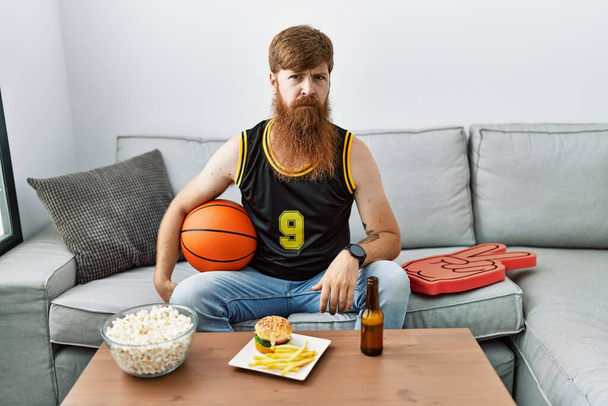 Homme caucasien à longue barbe tenant ballon de basket-ball acclamant jeu de télévision sceptique et nerveux, fronçant les sourcils bouleversés en raison de problèmes. personne négative.  - Photo, image