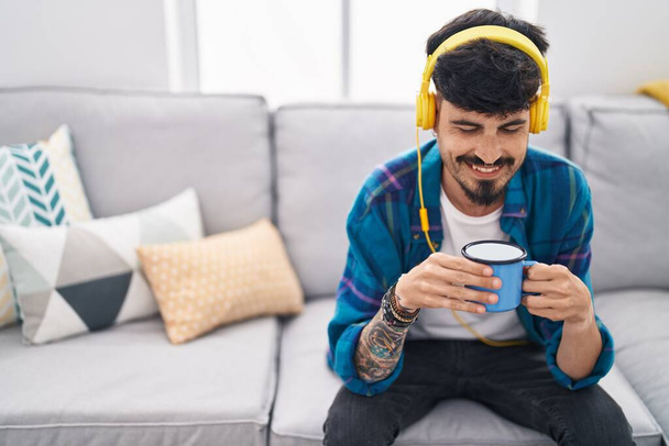 Young hispanic man listening to music drinking coffee at home - 写真・画像