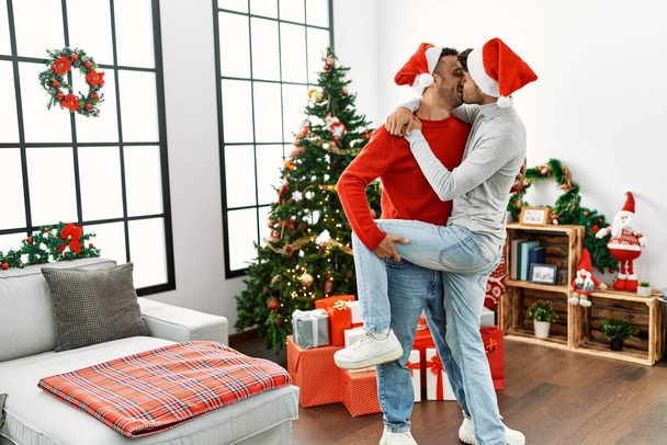 Two hispanic men couple kissing and dancing by christmas tree at home - Фото, изображение