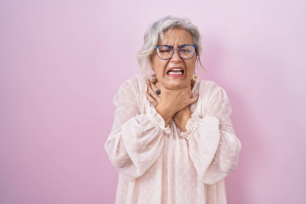 Frau mittleren Alters mit grauen Haaren steht vor rosa Hintergrund schreien und ersticken, weil qualvolle Würgen. Gesundheitsprobleme. Erstickungs- und Selbstmordkonzept.  - Foto, Bild