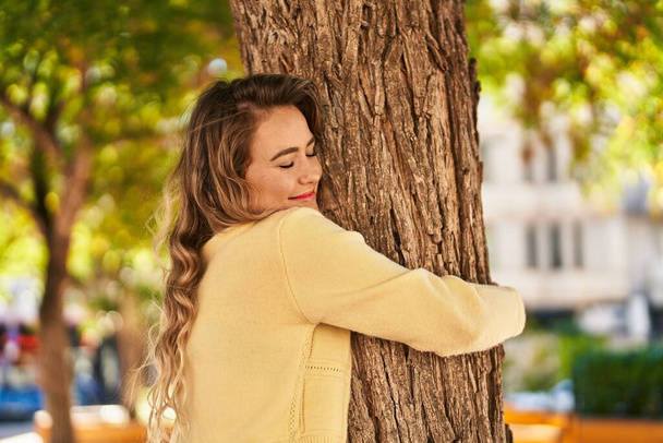 Молодая женщина улыбается уверенно обнимая дерево в парке - Фото, изображение