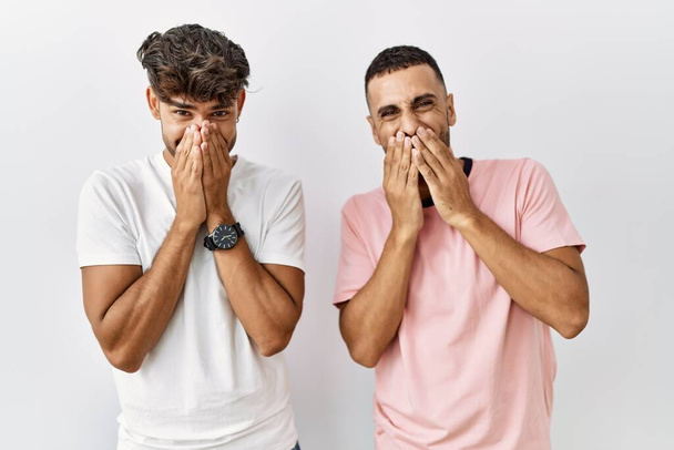 jong gay paar staande over geïsoleerde achtergrond lachen en beschaamd giechelen bedekking mond met handen, roddel en schandaal concept  - Foto, afbeelding