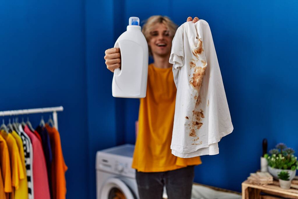 Jeune homme blond tenant t-shirt sale et détergent à la buanderie - Photo, image