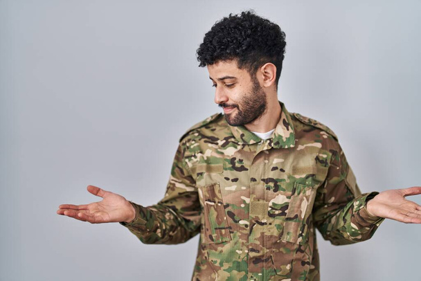 Arabe homme portant uniforme de camouflage de l'armée souriant montrant les deux mains paumes ouvertes, présentant et la publicité comparaison et l'équilibre  - Photo, image