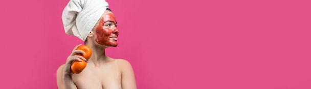 Szépségportré egy fehér törölközős nőről a fején piros tápláló maszkkal az arcán. Bőrápolás öko-organikus kozmetikai spa relaxációs koncepció. Egy lány áll a hátával egy narancssárga mandarinnal a kezében.. - Fotó, kép