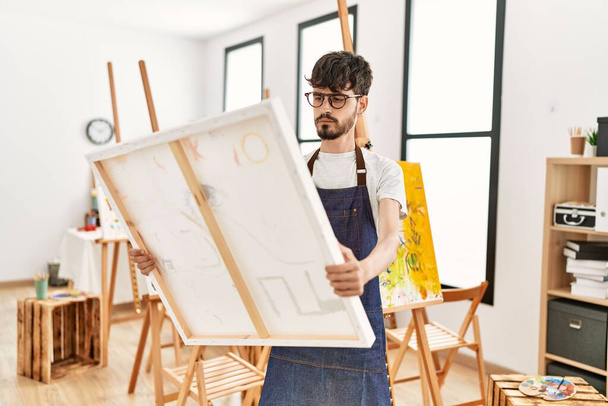 Young hispanic artist man concentrate holding canvas at art studio. - 写真・画像