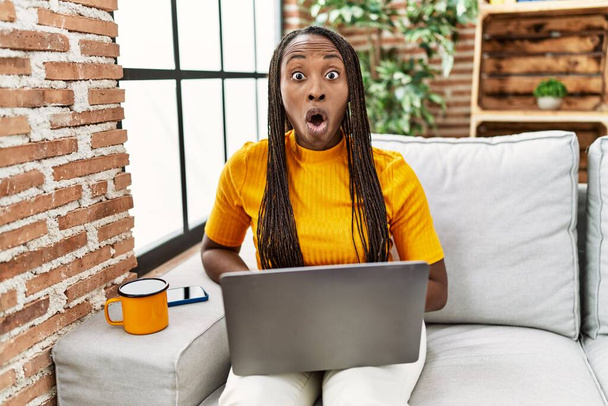 Африканская женщина сидит на диване, используя ноутбук дома испуганный и удивленный с открытым ртом для удивления, неверие лицо  - Фото, изображение