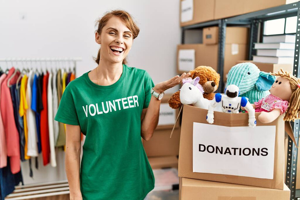 Schöne kaukasische Frau im freiwilligen T-Shirt am Spendenstand lächelt fröhlich und zeigt mit der Handfläche in die Kamera.  - Foto, Bild