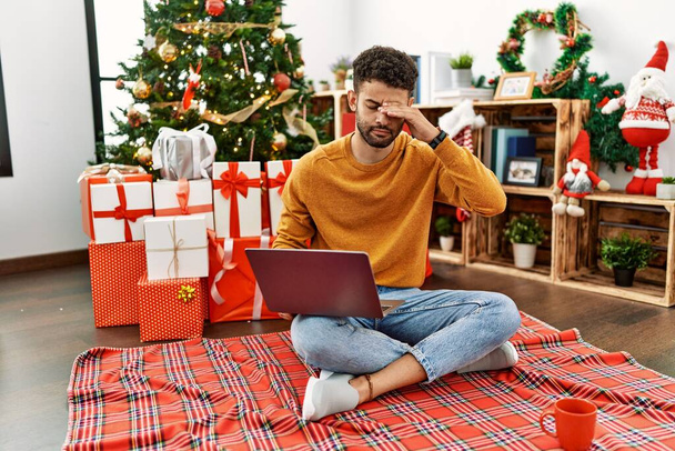 Arab young man using laptop sitting by christmas tree worried and stressed about a problem with hand on forehead, nervous and anxious for crisis  - 写真・画像