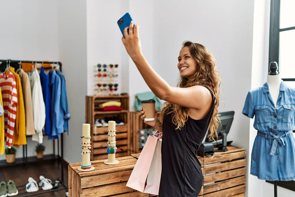 若いヒスパニック系のお客様の女性笑顔幸せなショッピングバッグを保持し、服店でスマートフォンによって自画自画自賛. - 写真・画像