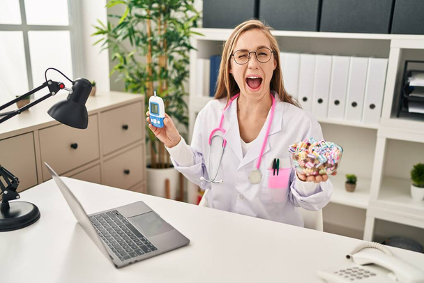 Jeune femme blonde médecin tenant glucomètre et bonbons clin d'oeil en regardant la caméra avec une expression sexy, gai et heureux visage.  - Photo, image