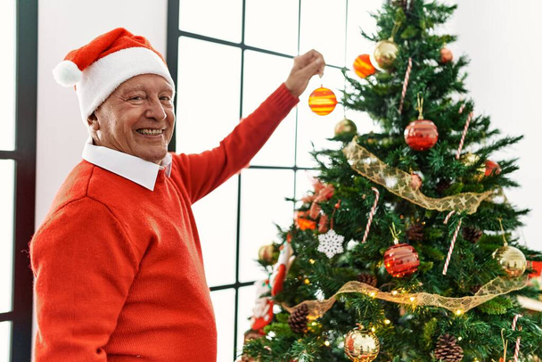 Ηλικιωμένος γκριζομάλλης χαμογελαστός χαρούμενος στολίζοντας χριστουγεννιάτικο δέντρο στο σπίτι. - Φωτογραφία, εικόνα