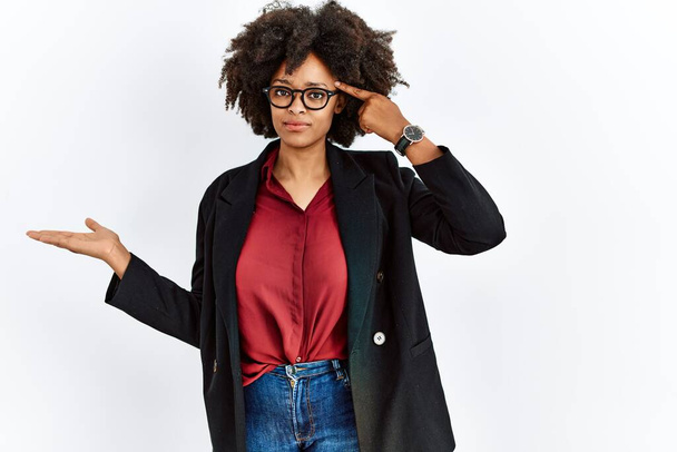 Mujer afroamericana con pelo afro vistiendo chaqueta de negocios y gafas confundidas y molestas con la palma abierta mostrando espacio para copiar y señalando con el dedo a la frente. Piénsalo.  - Foto, Imagen