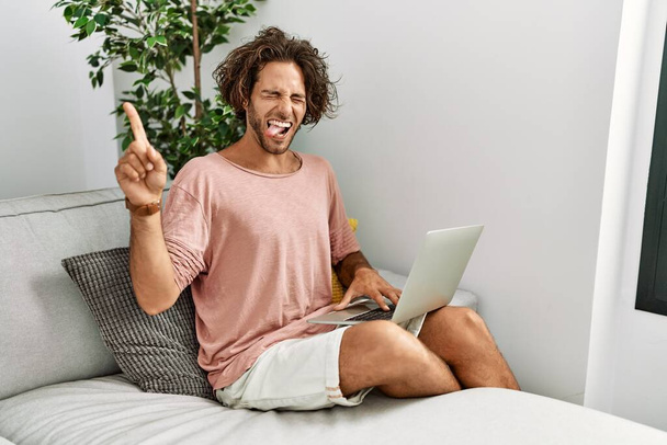 Junger hispanischer Mann, der zu Hause auf dem Sofa sitzt und mit Laptop die Zunge herausstreckt, glücklich mit lustigem Gesichtsausdruck. Emotionales Konzept.  - Foto, Bild