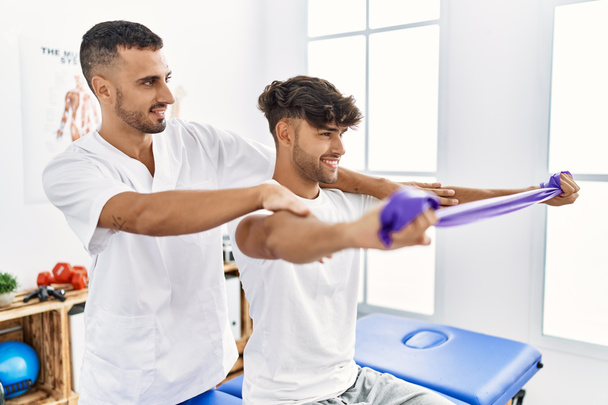 Двоє іспаномовних чоловіків фізіотерапевт і пацієнт мають сеанс реабілітації за допомогою еластичної смуги в клініці
 - Фото, зображення