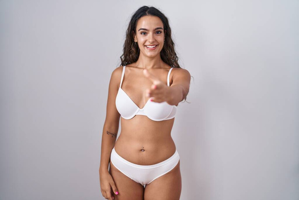 Молодая испаноязычная женщина в белом белье улыбается дружелюбно предлагая рукопожатие в качестве приветствия и приветствия. успешный бизнес.  - Фото, изображение