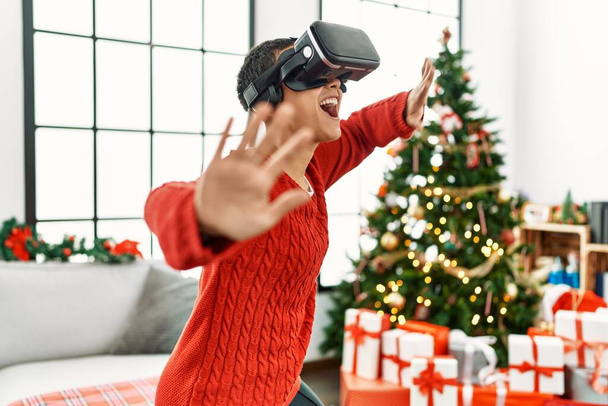 Junge hispanische Frau spielt Videospiel mit Vr-Brille, die zu Hause am Weihnachtsbaum steht - Foto, Bild