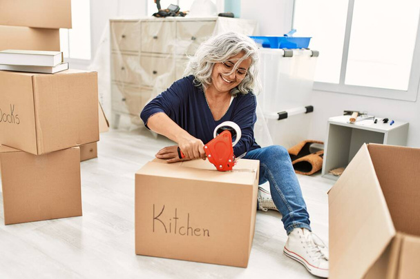 Keski-ikäinen harmaa tukka nainen hymyilee onnellinen pakkaus keittiö pahvilaatikko uudessa kodissa. - Valokuva, kuva
