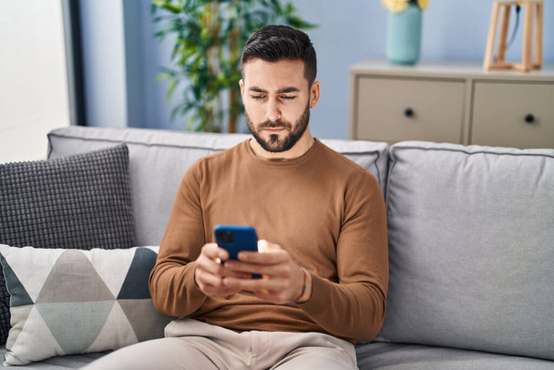 Giovane uomo ispanico utilizzando smartphone seduto sul divano a casa - Foto, immagini
