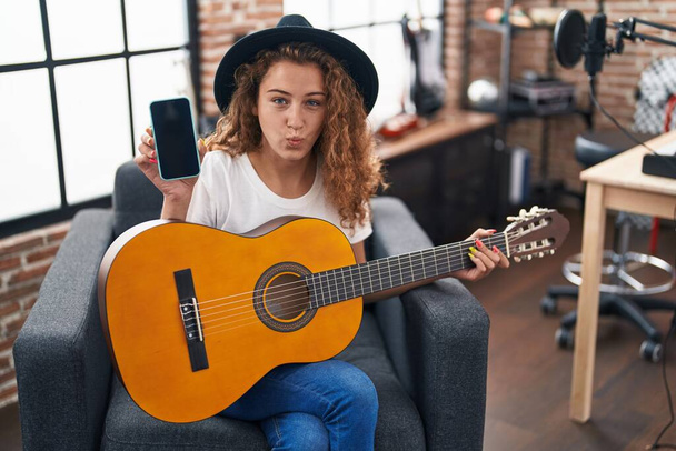 Молодая белая женщина, играющая на классической гитаре в музыкальной студии, держит смартфон, пыхтящий щеками со смешным лицом. рот надутый воздухом, ловящий воздух.  - Фото, изображение