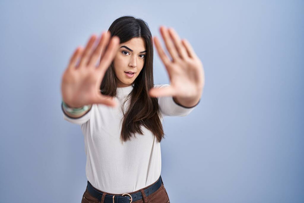 Young brunette woman standing over blue background doing frame using hands palms and fingers, camera perspective  - Photo, Image