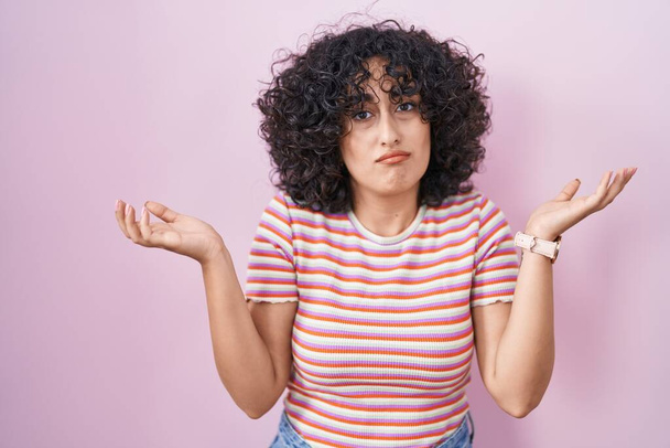 Young middle east woman standing over pink background clueless and confused expression with arms and hands raised. doubt concept.  - Foto, Imagem