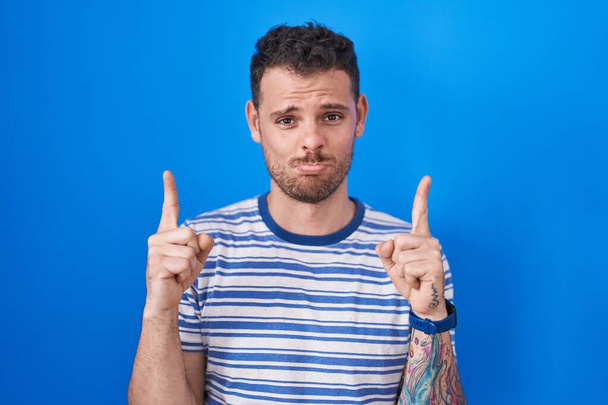Junger hispanischer Mann, der vor blauem Hintergrund steht und traurig und aufgebracht nach oben zeigt, mit den Fingern die Richtung anzeigt, unglücklich und deprimiert.  - Foto, Bild