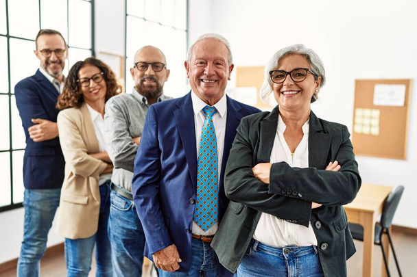 Группа бизнесменов среднего возраста улыбается счастливой стоя со скрещенными руками жестом в офисе - Фото, изображение