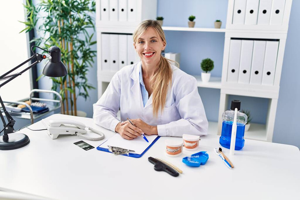 Young blonde woman wearing dentist uniform sitting on table at clinic - 写真・画像