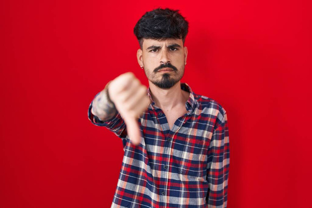 Homem hispânico jovem com barba em pé sobre fundo vermelho olhando infeliz e irritado mostrando rejeição e negativo com polegares para baixo gesto. má expressão.  - Foto, Imagem
