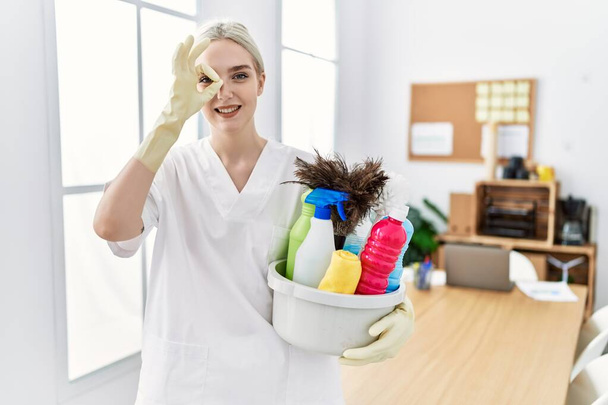 Молода біла жінка в чистішій формі тримає прибиральницю, усміхаючись, щасливо роблячи знак з рукою на око, дивлячись крізь пальці
  - Фото, зображення