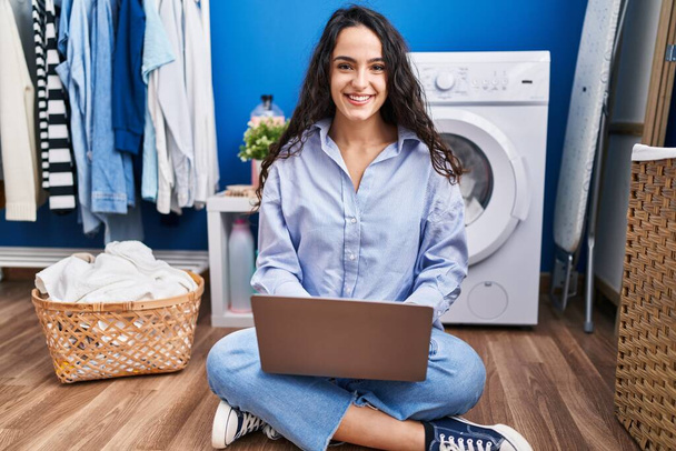 Молода іспаномовна жінка використовує ноутбук, який чекає на пральну машинку в пральні
 - Фото, зображення